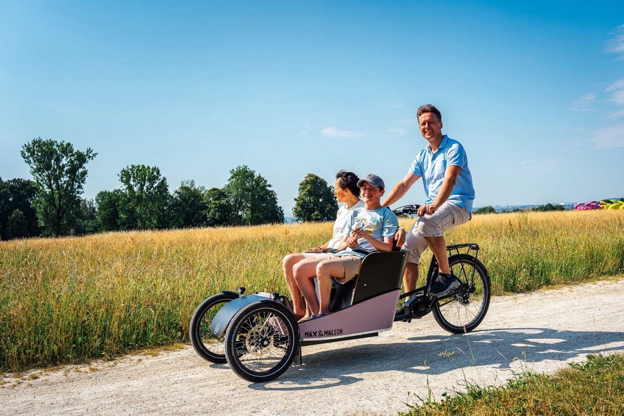 Vater fährt Partnerin und Sohn im Passenger Bike über einen Feldweg