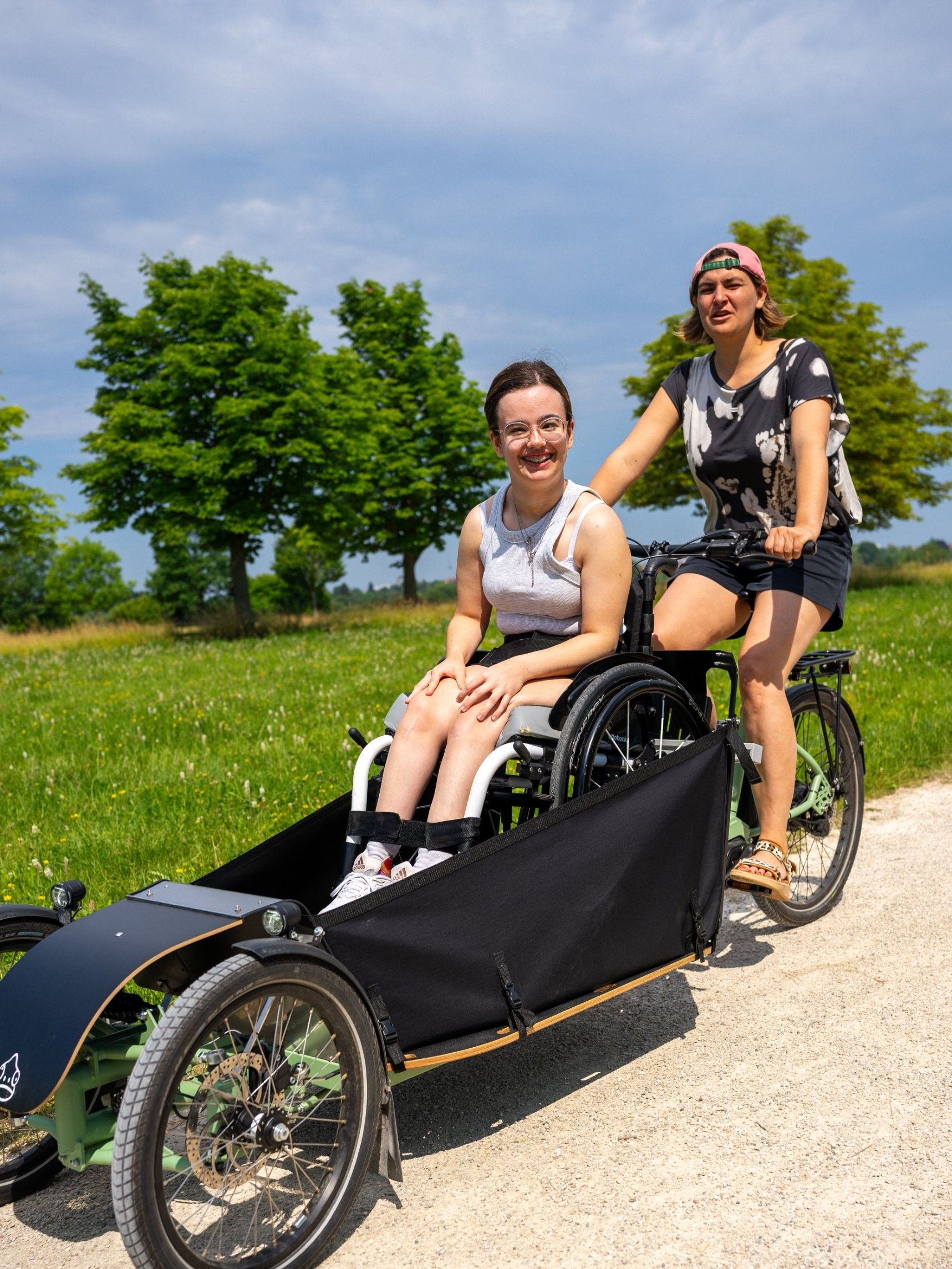 Frau fährt eine Rollstuhlfahrerin im Lastenrad durch einen Park